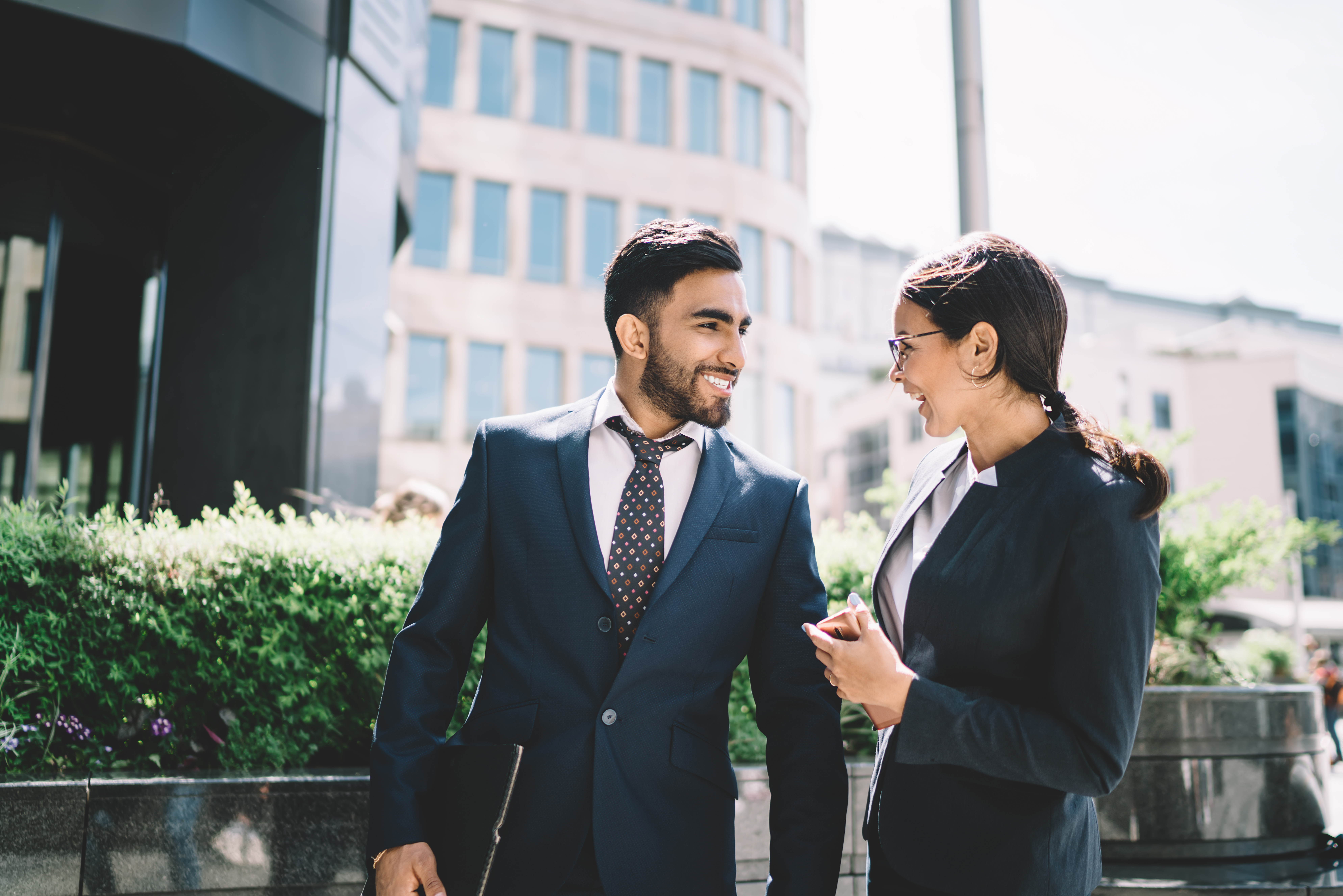 Successful smiling financial directors smiling outdoors during friendly conversation on break with office buildings on background, happy multicultural man and woman in formal wear communicate outdoors